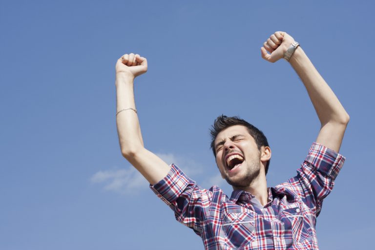 Purposeful man throws his arms in the air in celebration with blue sky in background