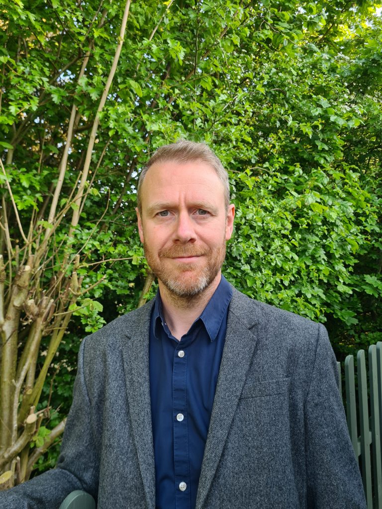 Jonny Baker, UK online integrative hypno-therapist for anxiety and life-meaning challenges, in front of a sage-coloured picket fence and a green tree, wearing a grey jacket and a blue shirt