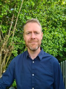 Jonny Baker, UK online integrative hypno-therapist for anxiety and life-meaning challenges, in front of a sage-coloured picket fence and a green tree, wearing a blue shirt